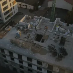 Wysoka perspektywa na plac budowy z widocznymi pracownikami w żółtych kamizelkach, szalunkami, zbrojeniami i częściowo wzniesionymi ścianami, z dźwigiem w tle.