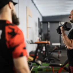 Mężczyzna ćwiczy unoszenie hantli w siłowni pod okiem drugiej osoby w koszulce z napisem 'TRENER'.