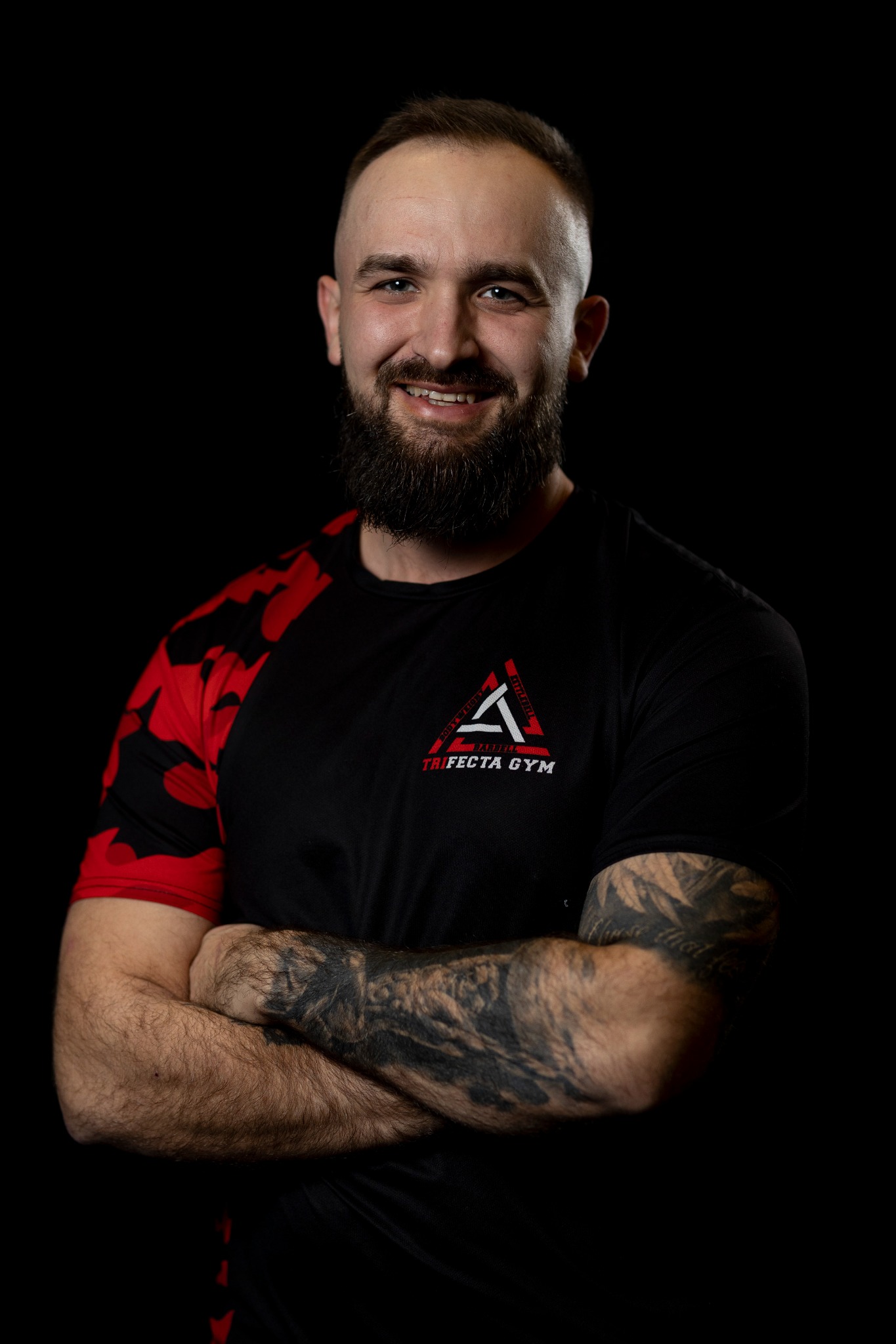 Uśmiechnięty mężczyzna z brodą i tatuażami na ramionach, ubrany w czarną koszulkę z logo Trifecta Gym na tle czarnego tła.