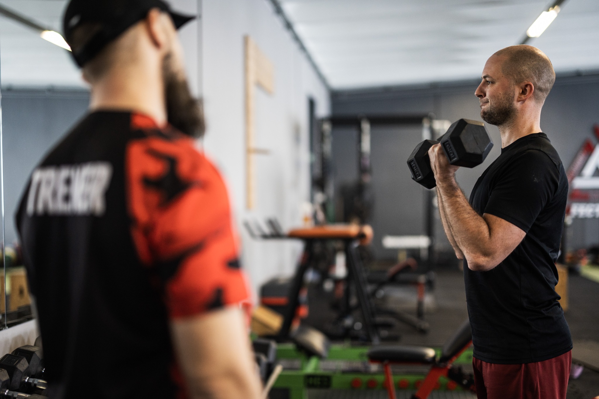Mężczyzna ćwiczy unoszenie hantli w siłowni pod okiem drugiej osoby w koszulce z napisem 'TRENER'.