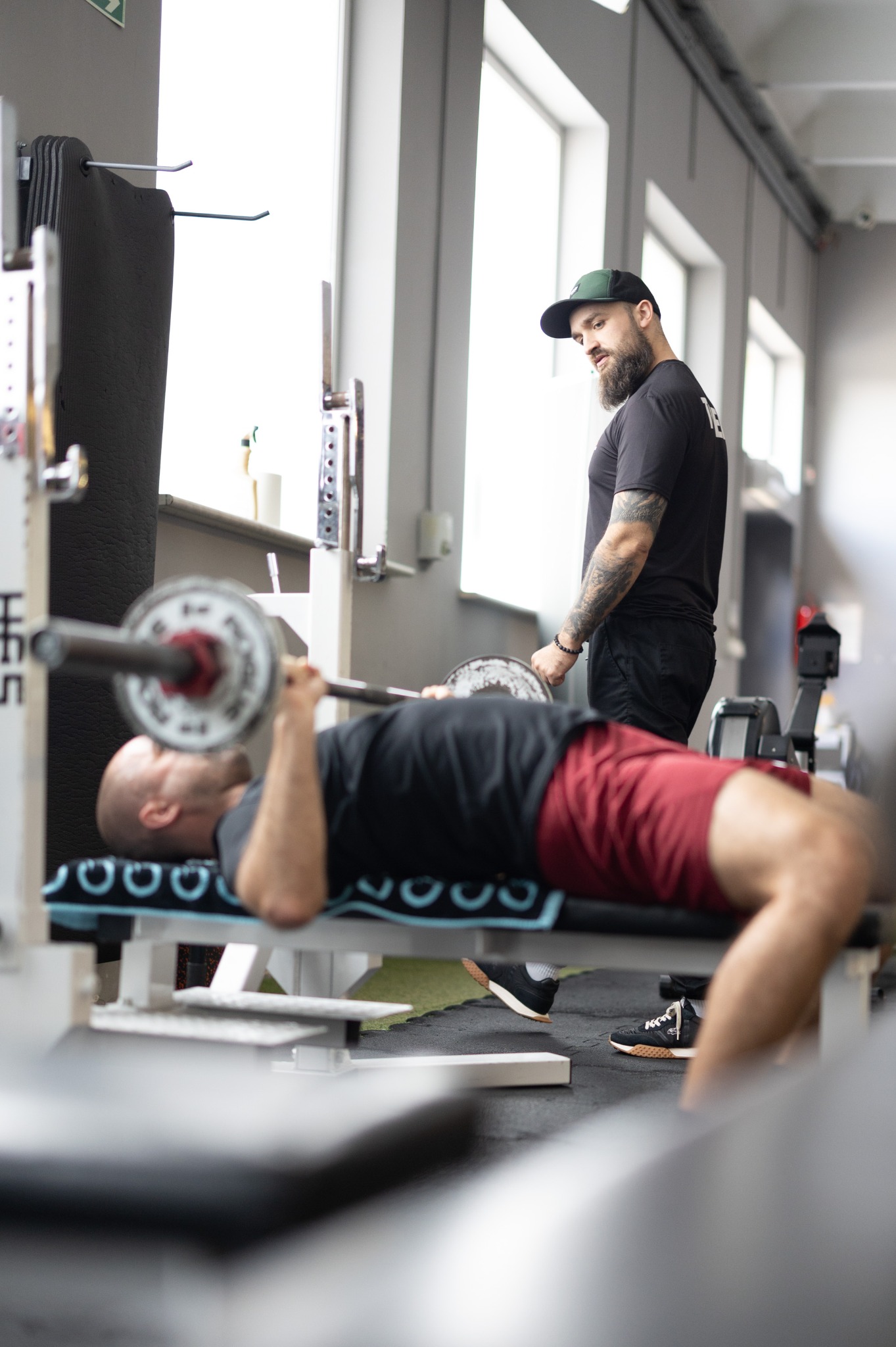 Mężczyzna ćwiczy wyciskanie sztangi na ławce, nadzorowany przez brodatego mężczyznę w czapce i z tatuażami na ramieniu w jasnej siłowni.