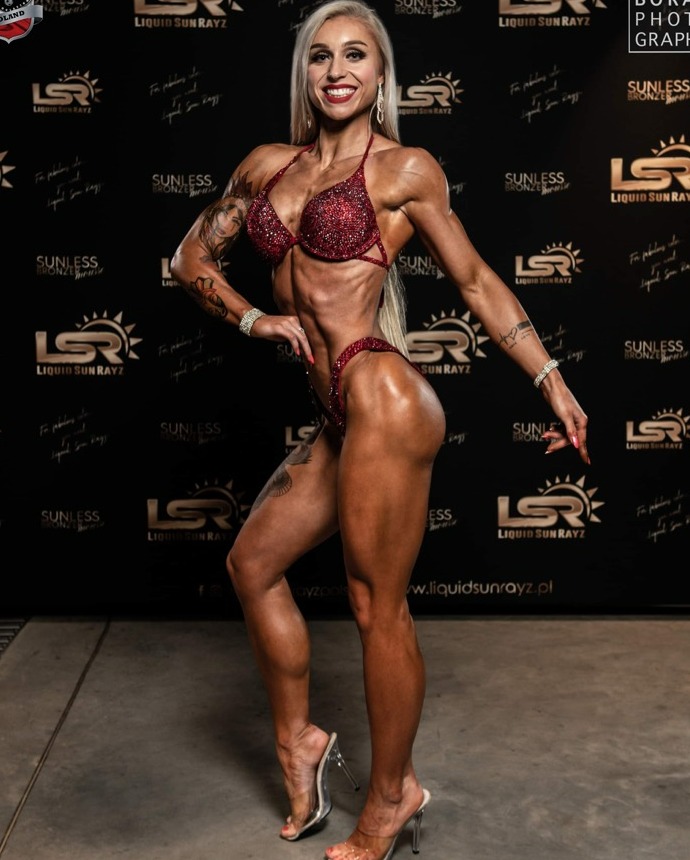 Uśmiechnięta kobieta w czerwonym, błyszczącym bikini prezentuje wyrzeźbioną sylwetkę na ciemnym tle z logotypami Liquid Sun Rayz.