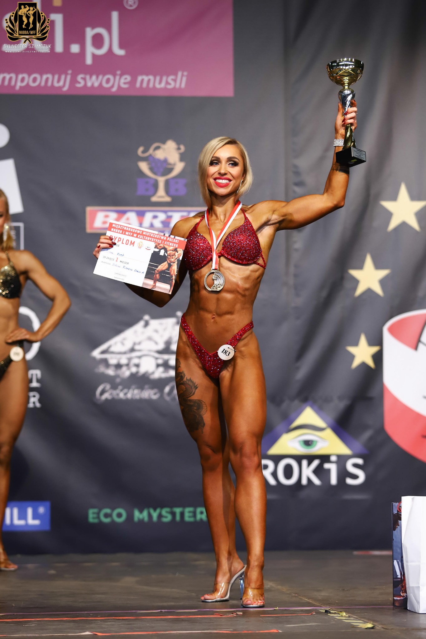 Kobieta w bikini fitness w czerwonych cekinach, z blond włosami, trzymająca puchar i dyplom za pierwsze miejsce na zawodach, na tle sceny z logo sponsorów.