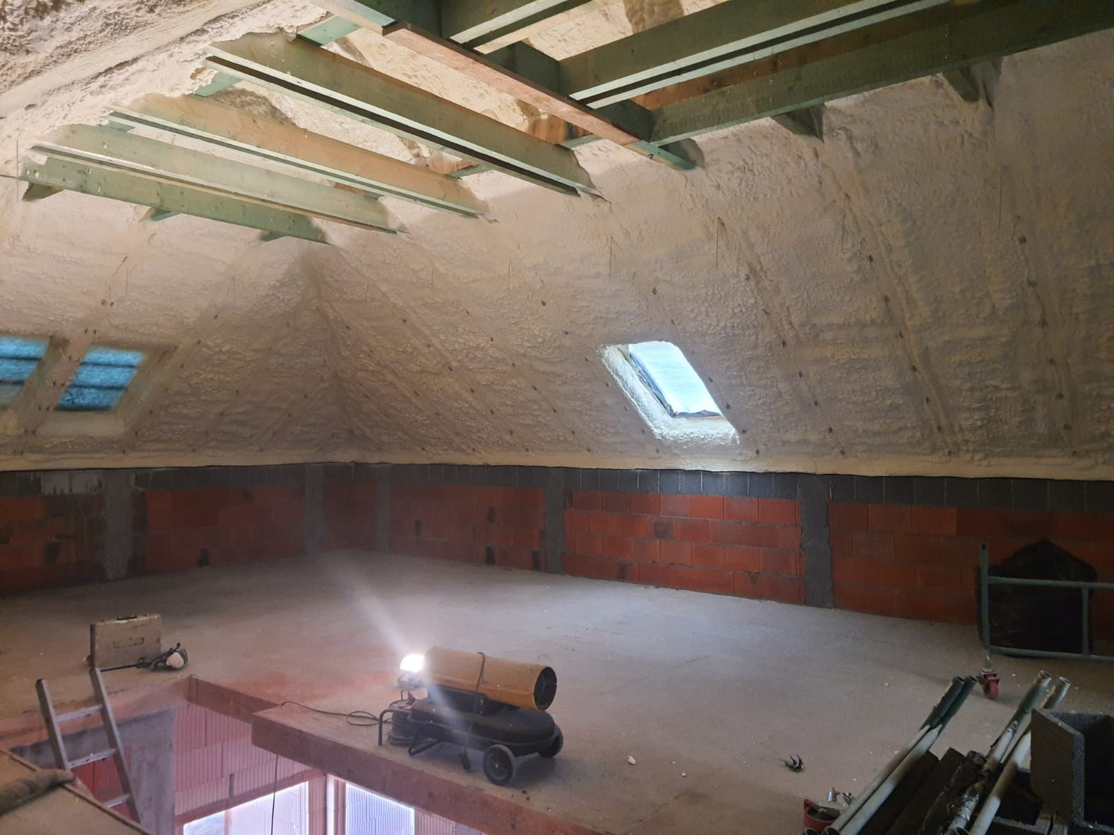 Poddasze po ociepleniu pianką PUR, widoczne krokwie i okno dachowe, ściany z czerwonej cegły, nagrzewnica na podłodze, oświetlenie robocze.