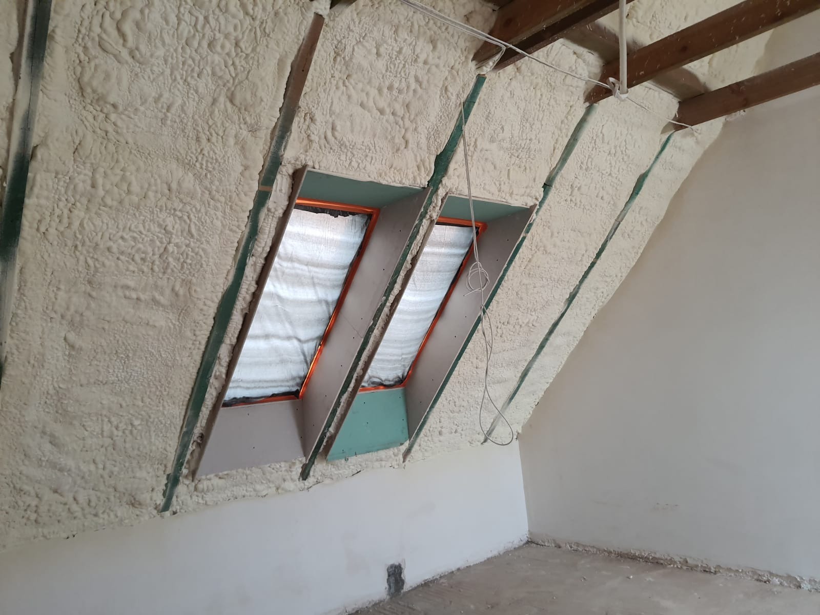 Poddasze z widoczną izolacją pianką PUR, dwa okna dachowe z folią odblaskową, drewniane elementy konstrukcyjne dachu, surowe ściany w trakcie wykańczania.