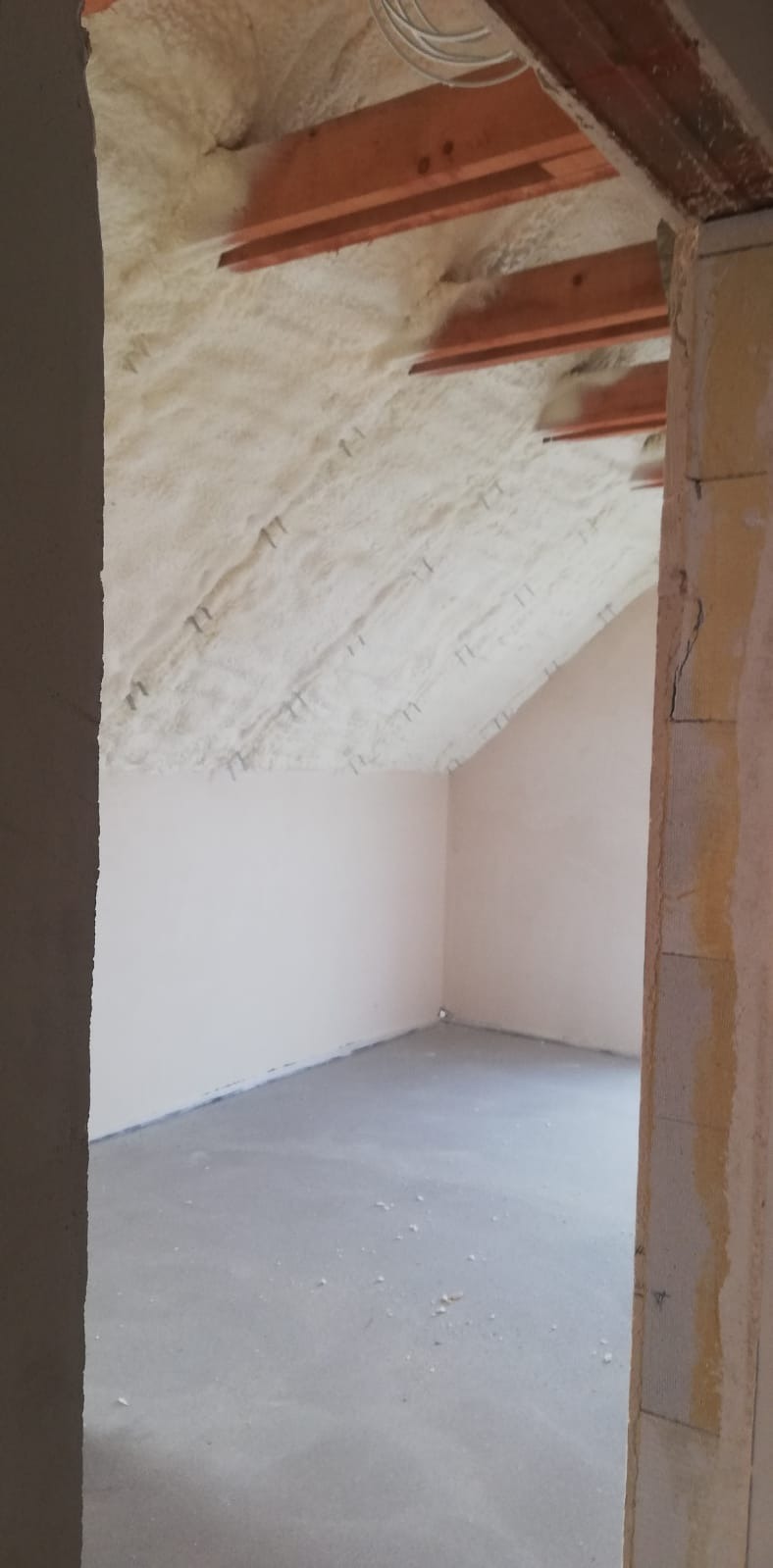 Poddasze ocieplone pianką, widoczne drewniane belki stropowe i szpilki mocujące izolację, surowe ściany i betonowa podłoga.