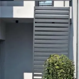 Nowoczesna, szara balustrada balkonowa z poziomych profili, uzupełniona pionowym panelem z tego samego materiału, przysłaniająca częściowo wejście do budynku, z niską roślinnością w tle.