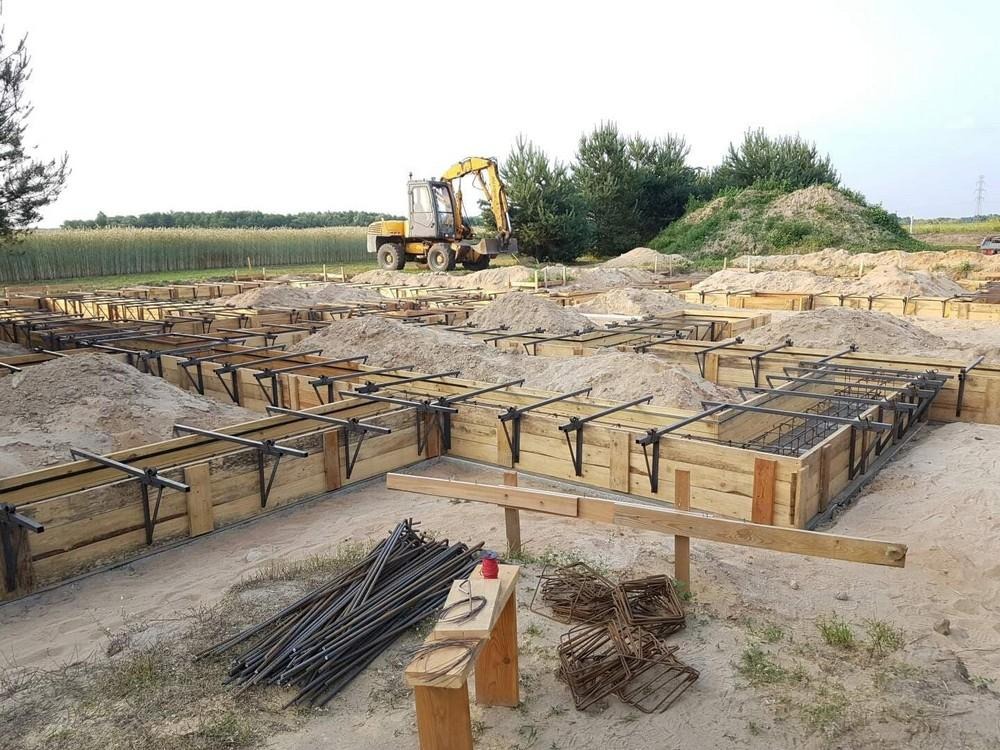 Przygotowanie fundamentów pod budynek: drewniane szalunki z metalowym zbrojeniem wypełnione piaskiem, w tle koparka i pole.