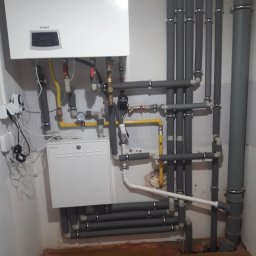 Usługi Hydrauliczne Łukasz Lewandowski - Kotłownia z białym piecem gazowym Termet, szarymi rurami izolowanymi termicznie i żółtymi rurami gazowymi, widoczne manometry i zawory.