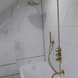 Nowoczesna łazienka z białą wanną i złotą armaturą: deszczownica, bateria wannowa z termostatem i wylewka, na tle płytek imitujących marmur z delikatnymi złotymi akcentami.