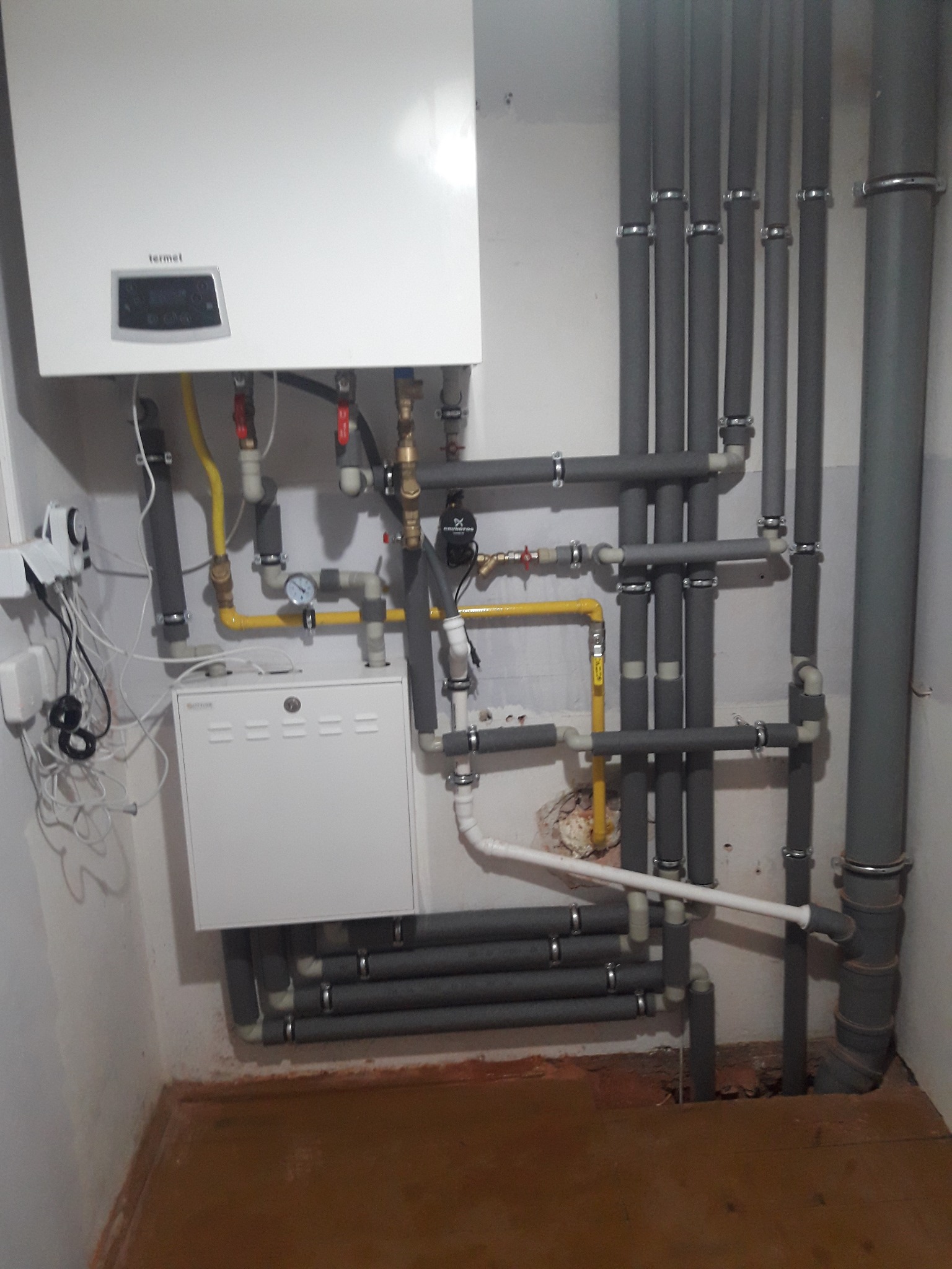 Kotłownia z białym piecem gazowym Termet, szarymi rurami izolowanymi termicznie i żółtymi rurami gazowymi, widoczne manometry i zawory.