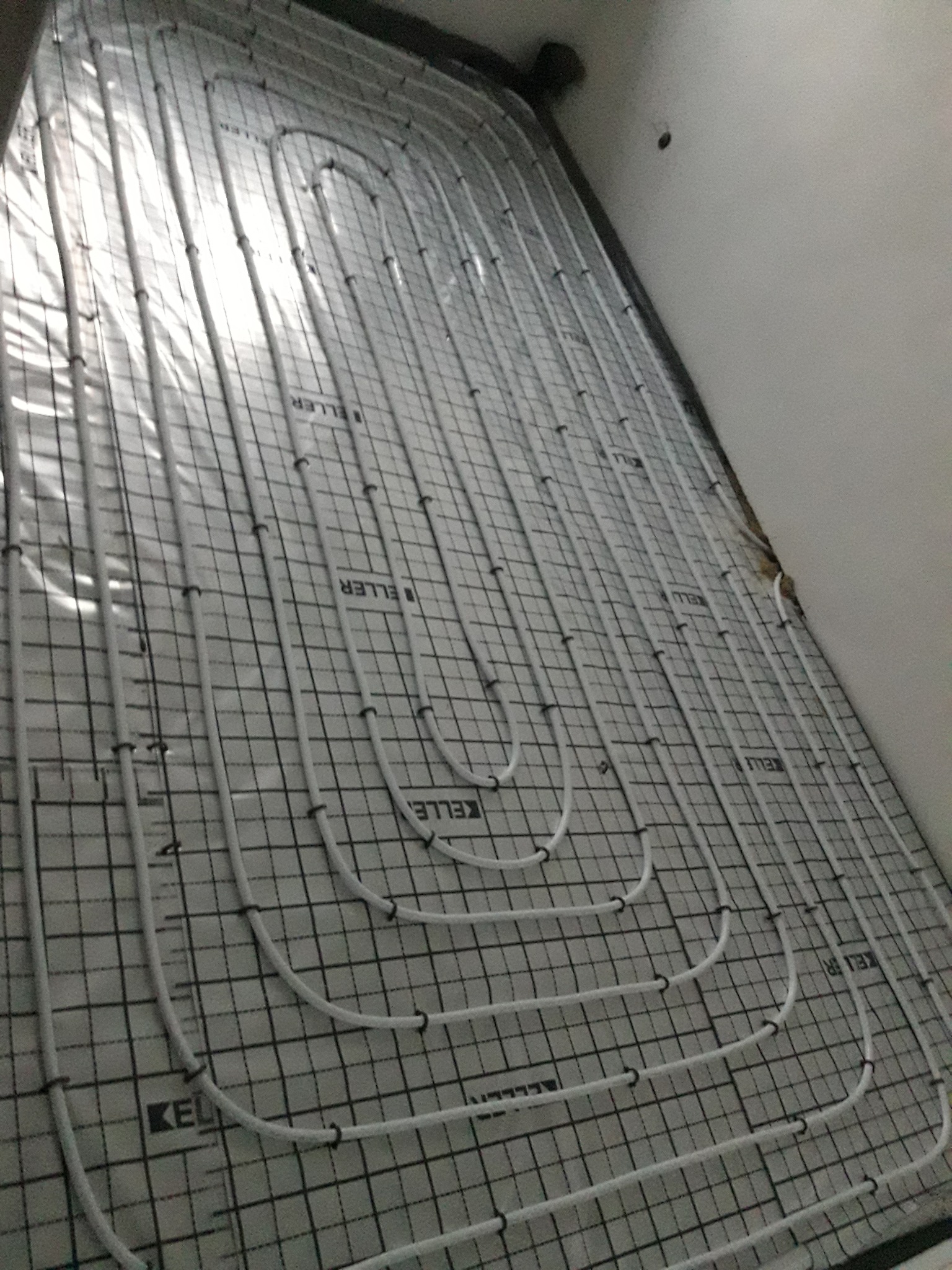 Białe rury systemu rozprowadzenia ciepła ułożone na siatce z tworzywa, przygotowane do zalania w posadzce.