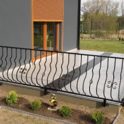 Czarna, metalowa balustrada balkonowa z dekoracyjnymi, kręconymi elementami, zamontowana na tarasie z szaro-białymi płytkami, widok z boku na tle szarej ściany domu i ogrodu.