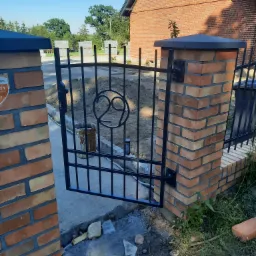 Otwarta, czarna, metalowa furtka z ozdobnym motywem w kształcie cyfry 29, osadzona na ceglanych słupkach z ciemnymi nakładkami, widoczna w trakcie montażu na posesji z ceglanym domem w tle.