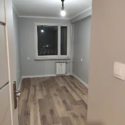 Pusty pokój po remoncie z panelami podłogowymi w odcieniach brązu, szare ściany, białe okno z widokiem na nocne miasto i grzejnik pod oknem, widoczna lampa wisząca z ozdobnym elementem.