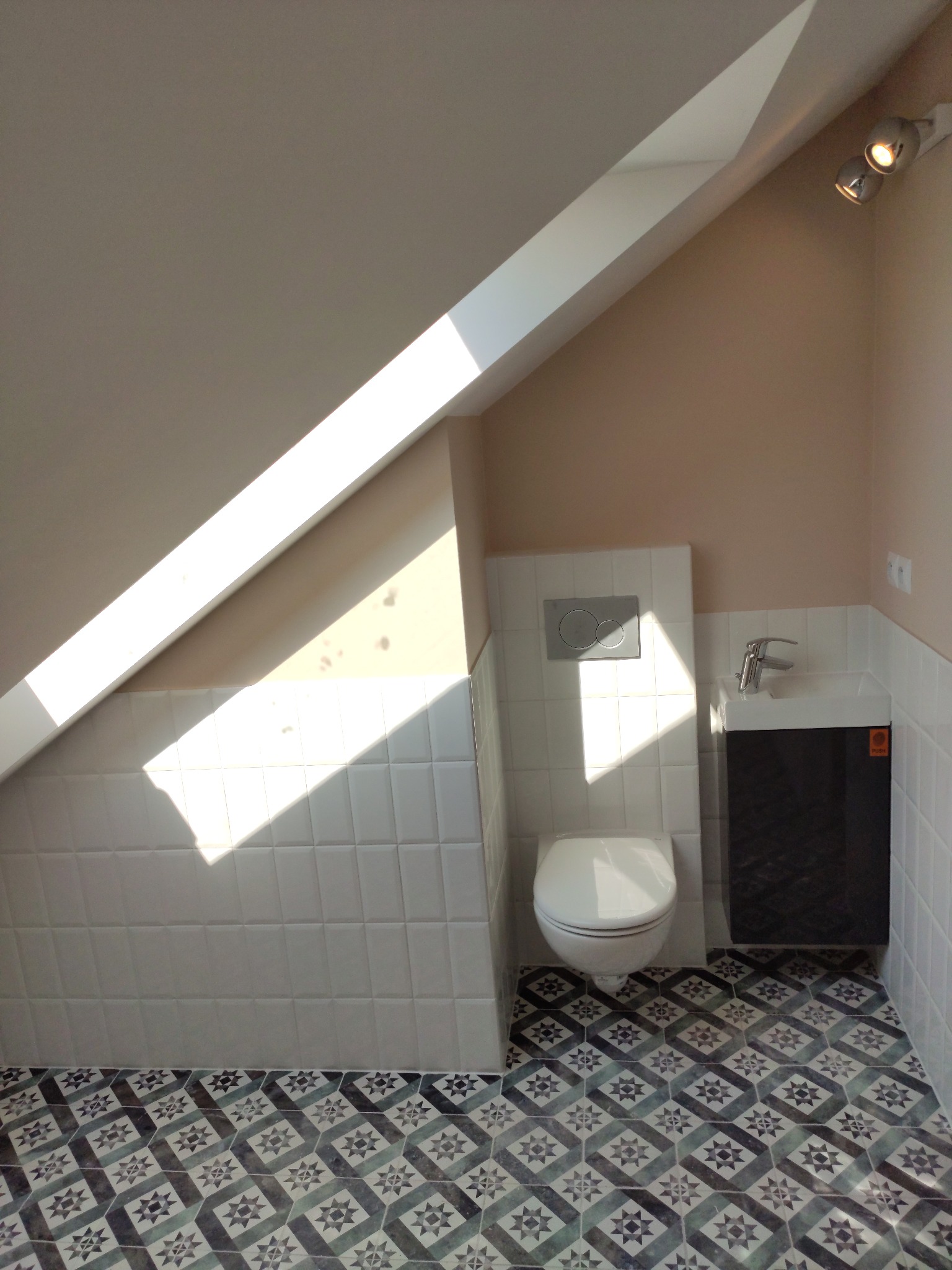 Nowoczesna łazienka na poddaszu z oknem dachowym, biała ceramika, umywalka z szafką w kolorze grafitowym i wzorzysta podłoga w odcieniach szarości i błękitu.