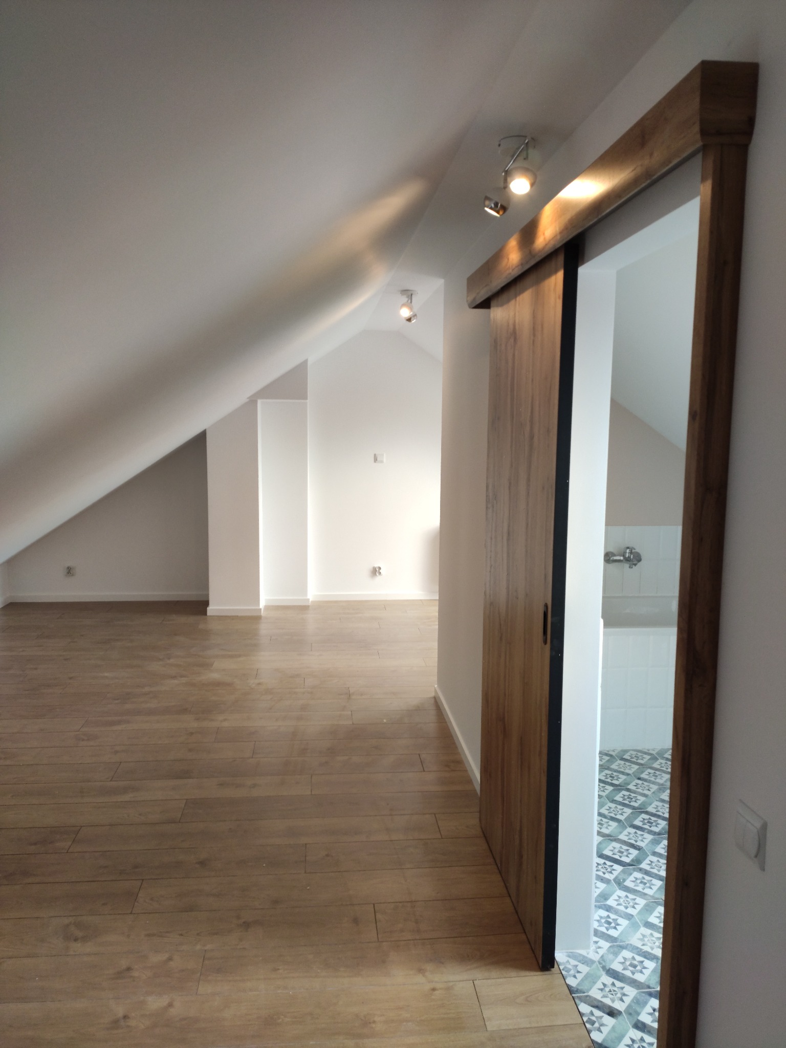 Poddasze z drewnianą podłogą i przesuwnymi drzwiami do łazienki z wzorzystą podłogą i białą armaturą.