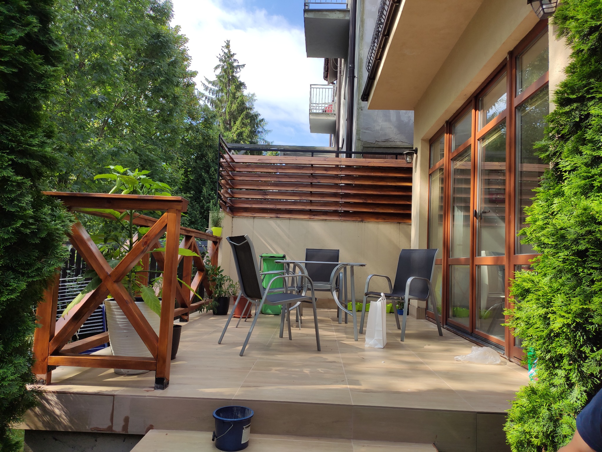 Zadaszony taras z drewnianą balustradą i brązowymi ramami okiennymi, wyposażony w metalowe krzesła i stolik, otoczony zielenią drzew i krzewów.