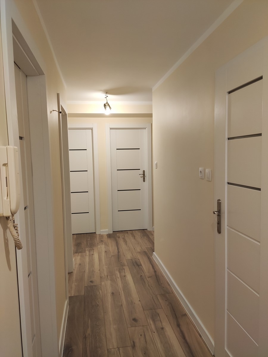 Korytarz po remoncie z panelami podłogowymi imitującymi drewno, kremowe ściany i białe drzwi z poziomymi, czarnymi akcentami. Widoczny biały domofon po lewej stronie.