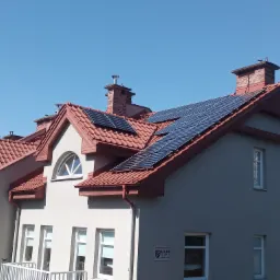 Panele słoneczne na dachu wielorodzinnego budynku z czerwoną dach&oacute;wką, widoczne kominy i jasne niebo.