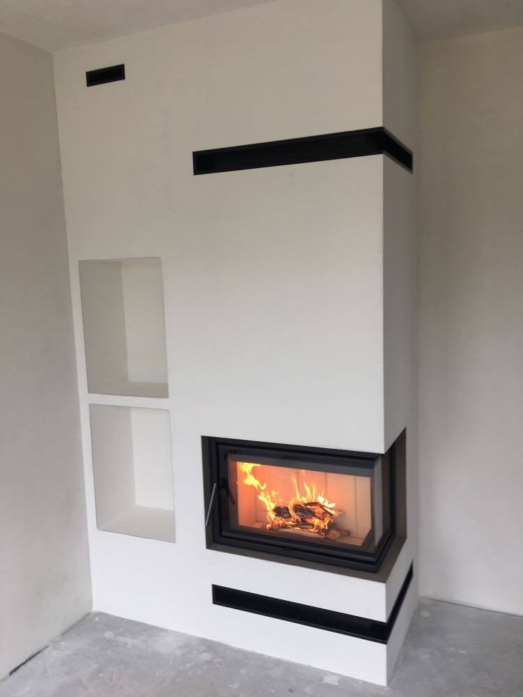 Nowoczesny narożny wkład kominkowy z palącym się drewnem, obudowany białą konstrukcją z czarnymi akcentami i wnękami po lewej stronie, widok z przodu.