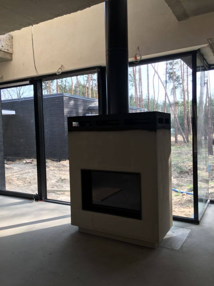 Surowy, minimalistyczny piec wolnostojący z czarnym kominem w nowoczesnym wnętrzu z dużymi oknami na tle lasu.