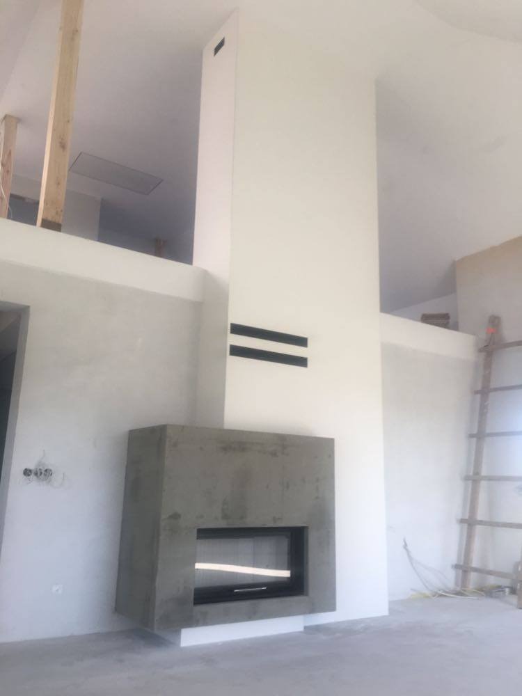 Surowy betonowy wkład kominkowy z czarną ramą, minimalistyczny design, widoczne elementy instalacji elektrycznej na ścianie, drabina w tle, wnętrze w trakcie wykończenia.