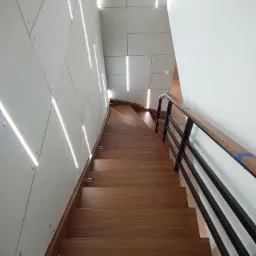 Wysoki kąt widzenia na drewniane schody z czarną balustradą i drewnianą poręczą, prowadzące w dół, z nowoczesnym oświetleniem LED na ścianie o geometrycznym wzorze.