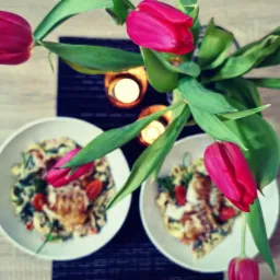 Dwa talerze z daniem makaronowym, udekorowane pomidorkami koktajlowymi i zielenią, otoczone świeżymi tulipanami i świecami, widok z góry.