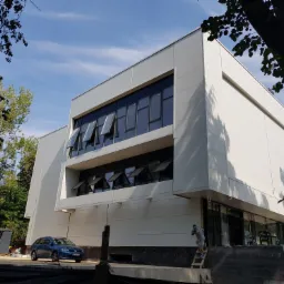 Nowoczesny budynek hali z białych płyt warstwowych, z otwartymi oknami, widziany z zewnątrz, z niebieskim samochodem i pracownikiem na drabinie, w słoneczny dzień.