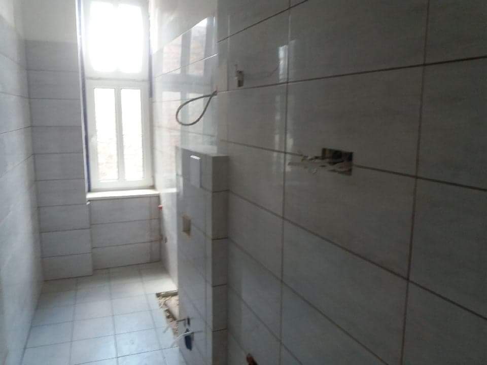 Wnętrze łazienki w trakcie prac, widoczne białe płytki na ścianach i podłodze, otwór na gniazdko elektryczne oraz wystające kable, okno z białymi ramami w tle.