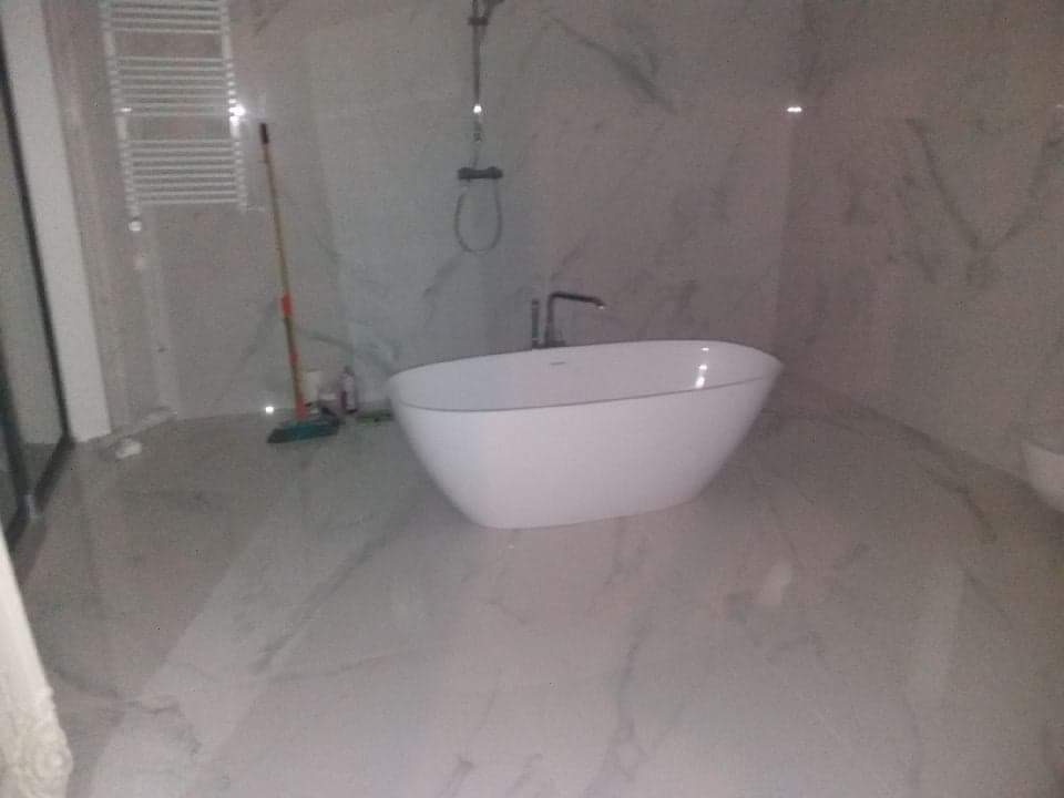 Nowoczesna, wolnostojąca wanna owalna w łazience wykończonej płytkami imitującymi marmur, widoczny grzejnik drabinkowy, bateria wannowa i częściowo mop do podłogi.