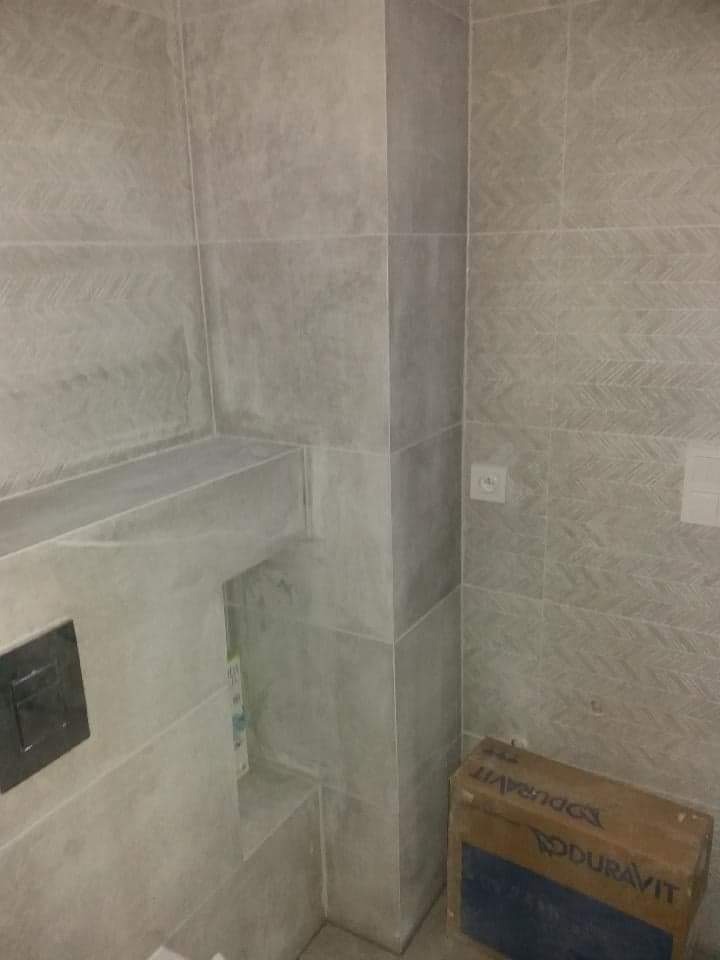 Wykończona łazienka z szarymi płytkami imitującymi beton, zabudowana półka i przycisk spłuczki w ścianie, karton po ceramice Duravit w rogu.