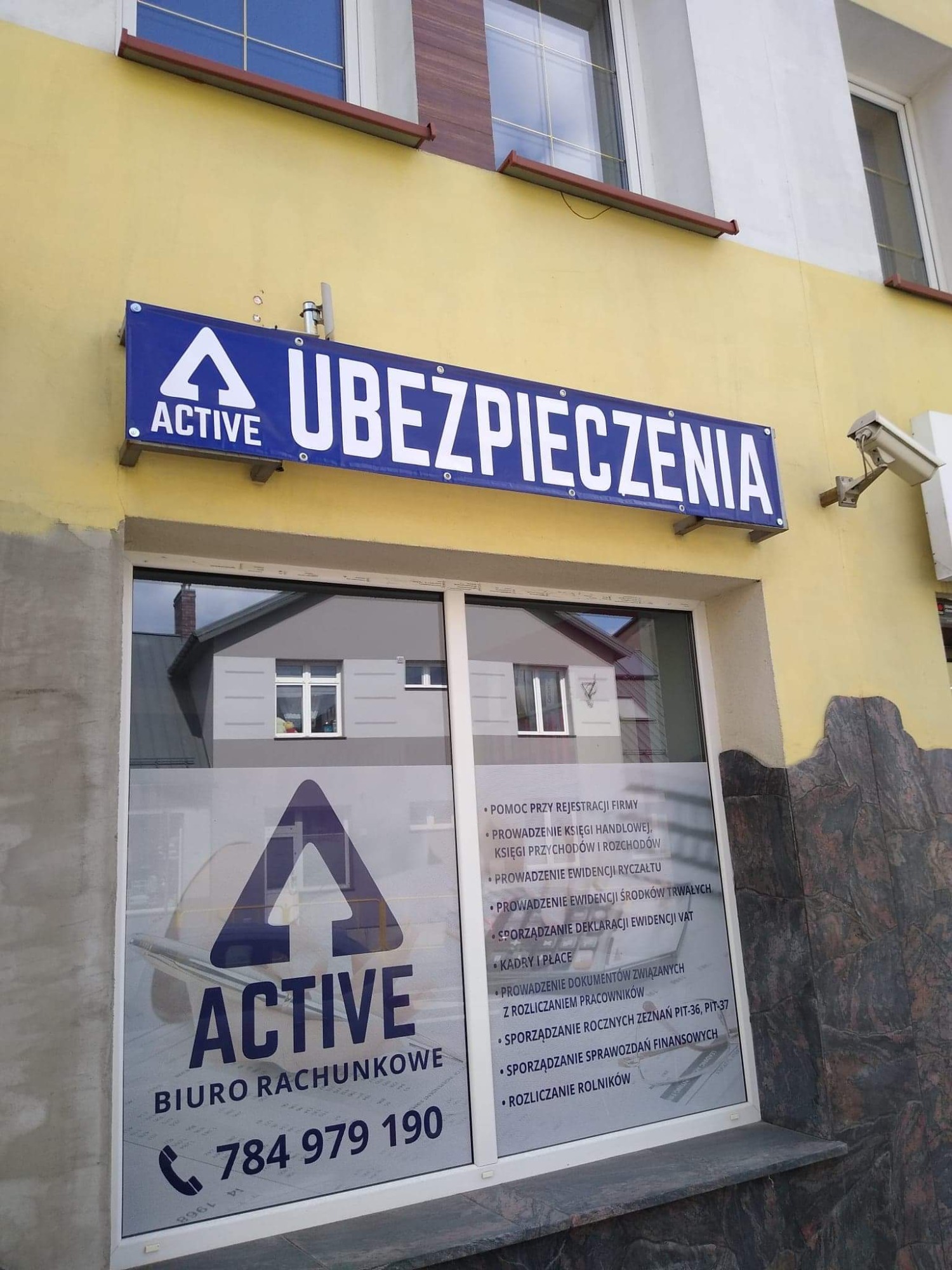 Fasada budynku z szyldem 'ACTIVE UBEZPIECZENIA' oraz naklejką na szybie 'BIURO RACHUNKOWE' z numerem telefonu, z widocznymi usługami księgowymi.