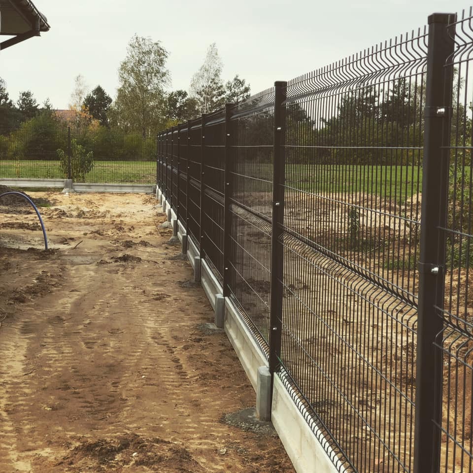 Nowo zainstalowane, brązowe ogrodzenie panelowe z podmurówką betonową na nierównym, piaszczystym terenie budowy, widok z boku.