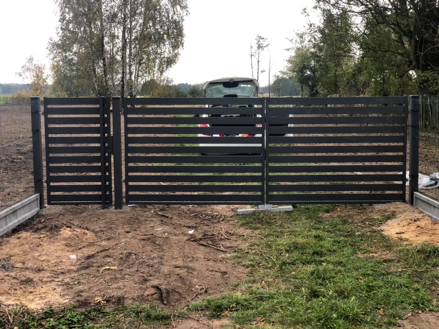 Nowoczesna, pozioma brama wjazdowa z czarnych profili stalowych, widok na otwarty bagażnik samochodu osobowego w tle, obok ogrodzenie z siatki.