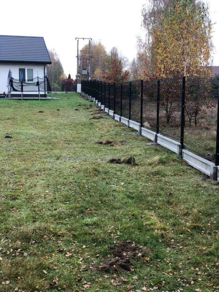 Czarny system ogrodzeniowy z paneli na betonowej podmurówce, widok z boku, z wykopanymi dołkami wzdłuż ogrodzenia, dom z trampoliną w tle.