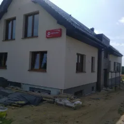 Dwukondygnacyjny dom z jasną elewacją i ciemną dachówką, z widocznym logo firmy budowlanej na ścianie, w trakcie prac wykończeniowych, z materiałami budowlanymi składowanymi wokół budynku.