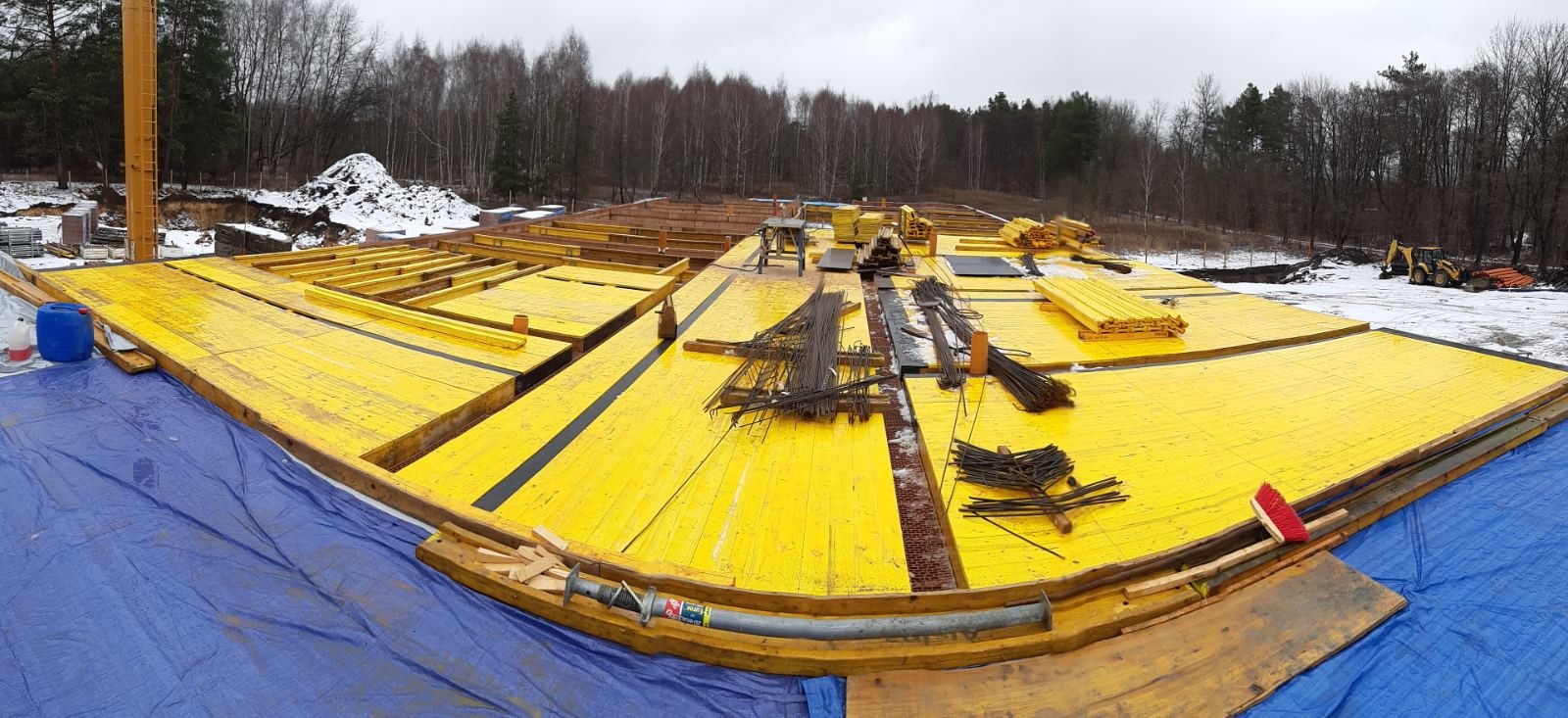 Szeroki kadr na żółte szalunki fundamentowe z widocznymi prętami zbrojeniowymi, przygotowane do zalania betonem, z koparką w tle i resztkami śniegu.