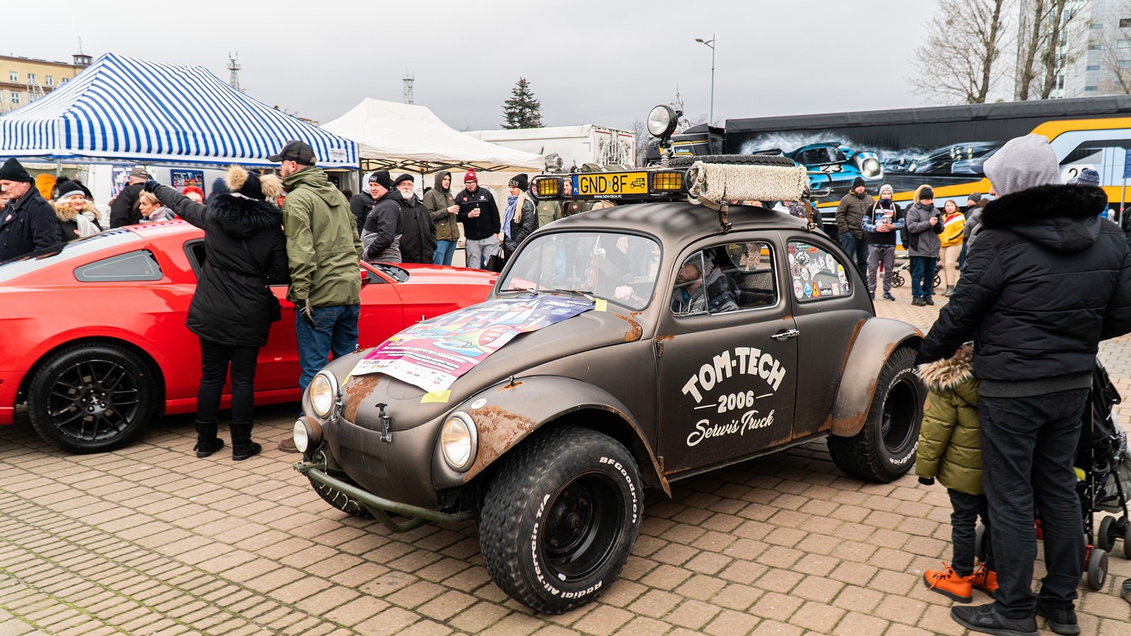 Zmodyfikowany, matowy Volkswagen Garbus z dużymi oponami terenowymi, lampą na dachu i naklejkami, stoi na brukowanym placu w otoczeniu ludzi i innych pojazdów, w tym czerwonego Forda Mustanga...