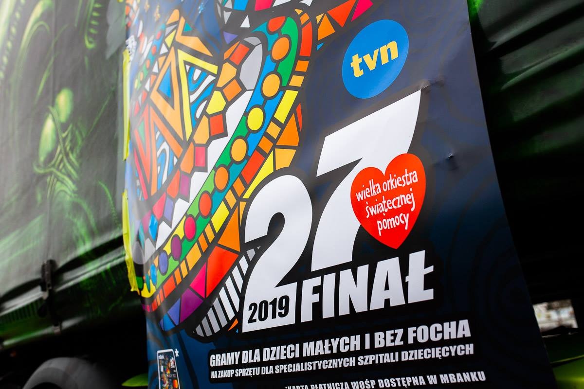 Plakat 27. Finału Wielkiej Orkiestry Świątecznej Pomocy na boku zielonego samochodu z grafiką obcego, z widocznym logo TVN.
