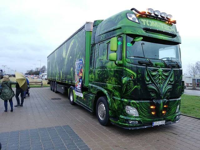 Zielona ciężarówka z naczepą, ozdobiona grafikami w stylu alien, stoi na parkingu z przechodniami w tle.