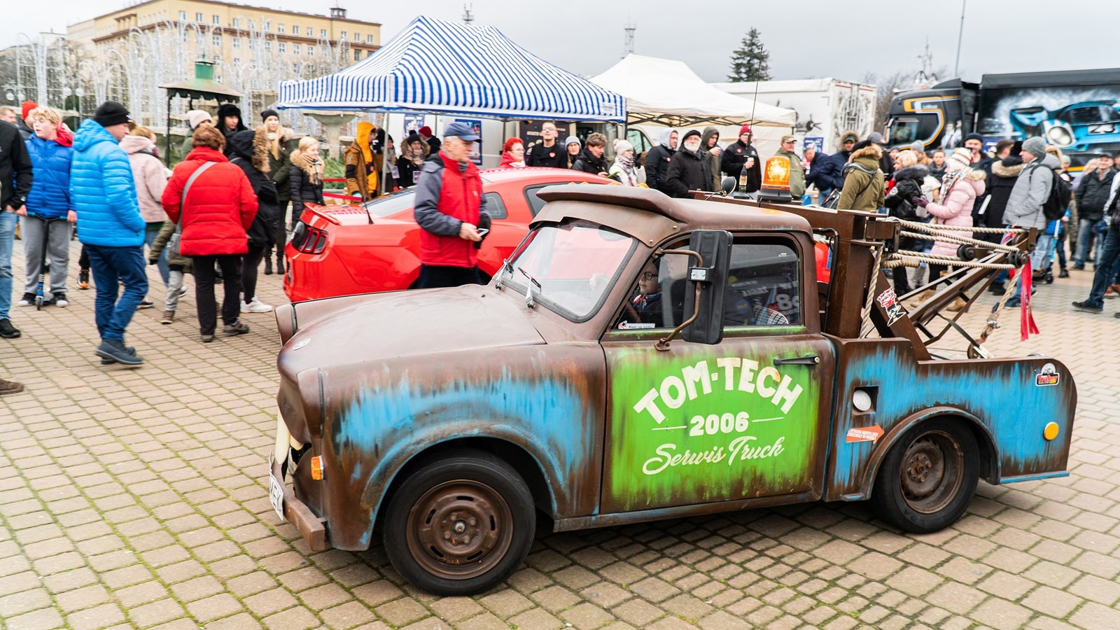 Zardzewiały, stylizowany na pojazd pomocy drogowej, samochód marki Trabant z napisem 'TOM-TECH 2006 Serwis Truck' na tle tłumu ludzi i czerwonego auta.