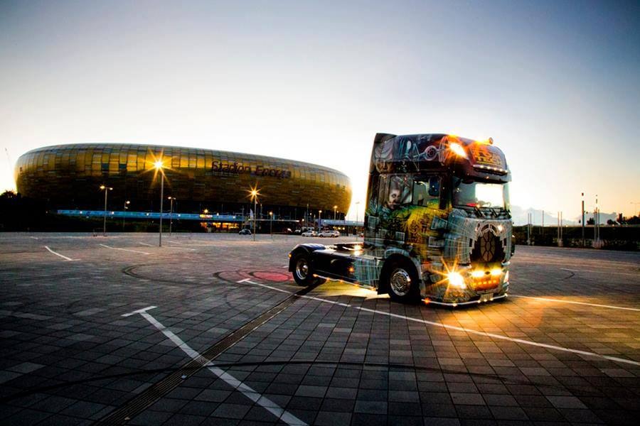 Artystycznie pomalowana ciężarówka z włączonymi światłami na tle Stadionu Energa Gdańsk o zmierzchu.