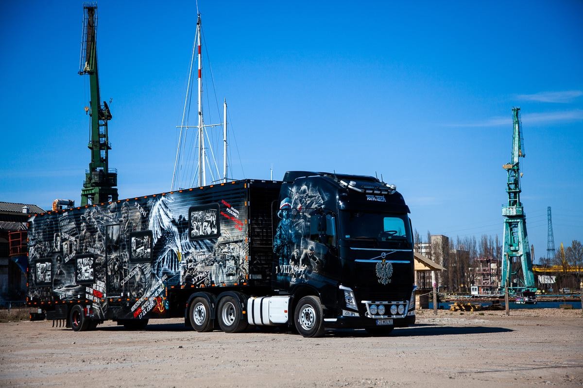 Czarna ciężarówka Volvo z naczepą ozdobiona patriotycznymi grafikami, stojąca na placu z dźwigami portowymi w tle.