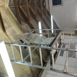 Pracownik w kombinezonie ochronnym, stojący na aluminiowej drabinie, aplikuje pianę izolacyjną na poddaszu z widocznymi drewnianymi elementami konstrukcyjnymi dachu.