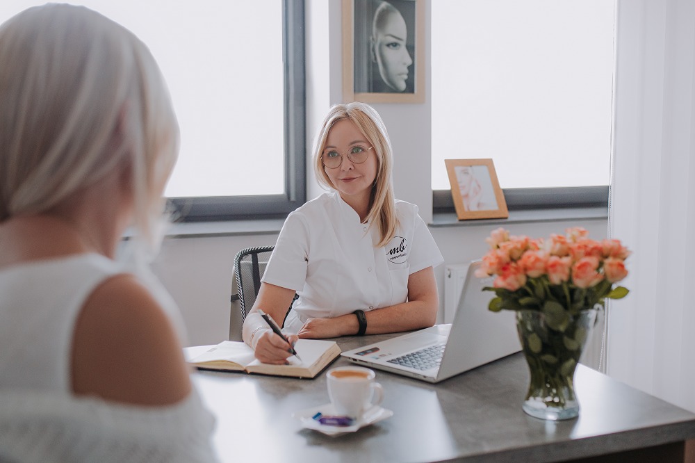 Konsultacja w gabinecie: blondynka w okularach, w białym uniformie, notuje w notesie przy biurku z laptopem, kawą i kwiatami. W tle portret kobiety.
