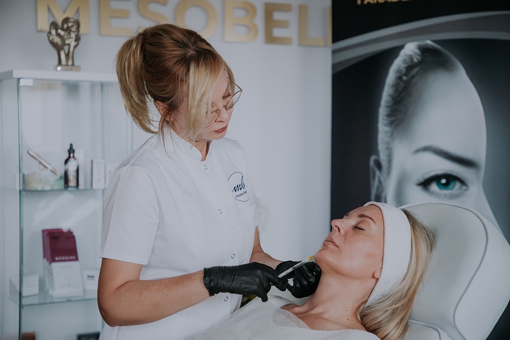 Zabieg mezoterapii igłowej szyi wykonywany przez kosmetologa w czarnych rękawiczkach. Klientka leży na fotelu zabiegowym z opaską na włosach.