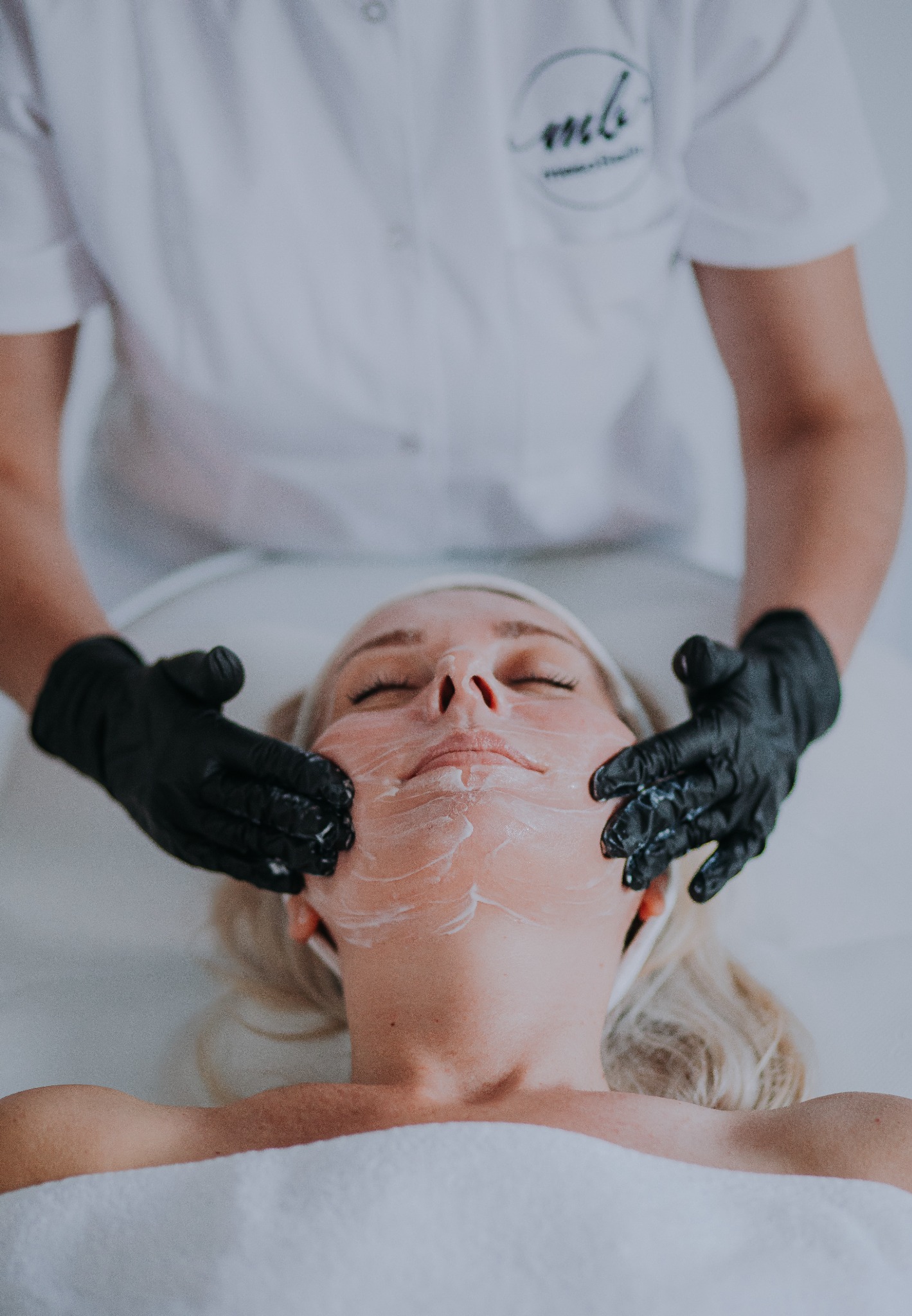 Zabieg kosmetyczny na twarz klientki wykonywany przez kosmetologa w czarnych rękawiczkach. Widoczny biały krem na skórze i logo na uniformie.