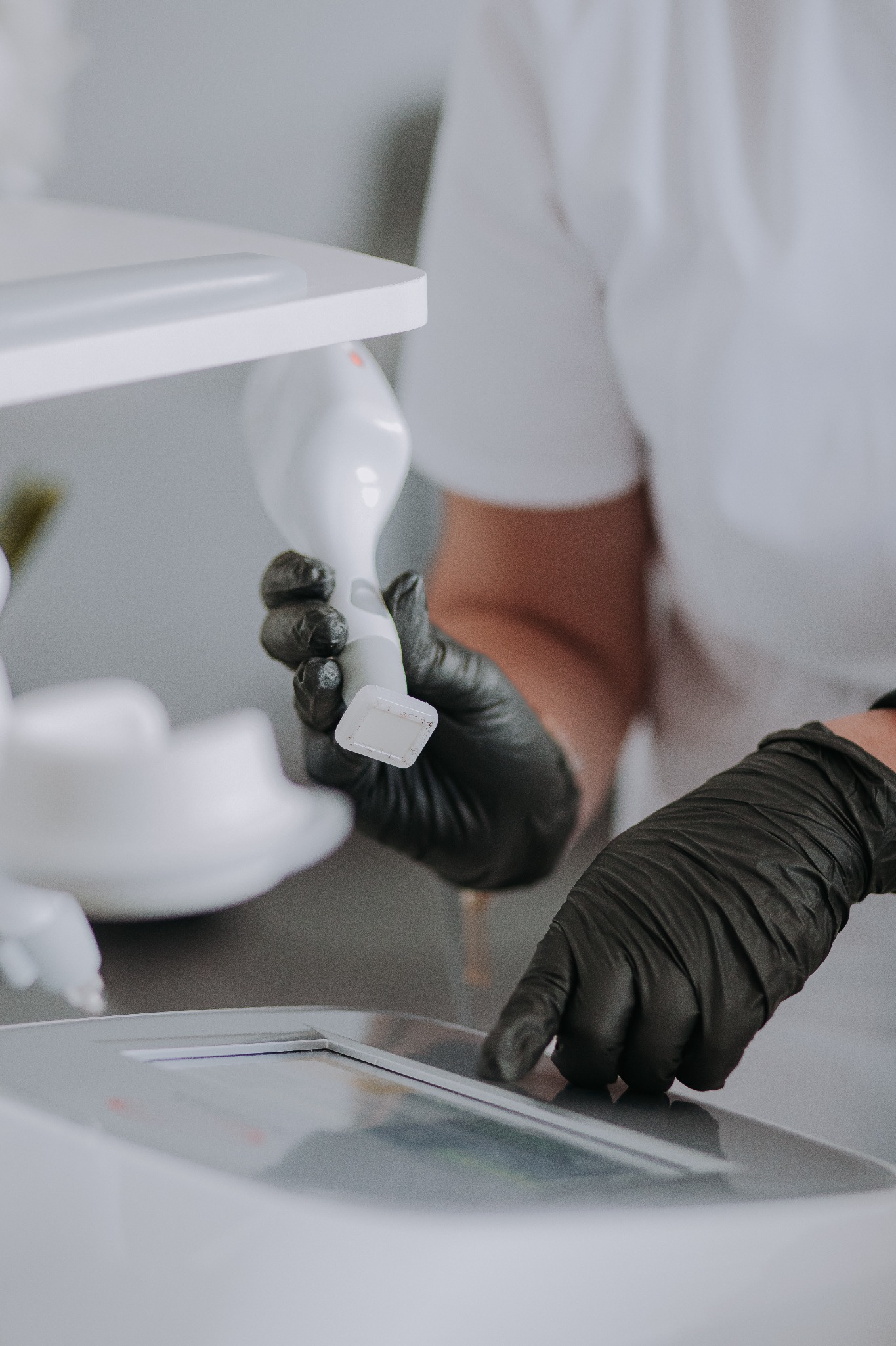 Obsługa urządzenia do zabiegów kosmetycznych w rękawicach ochronnych. Białe urządzenie, zbliżenie na dłonie i panel sterowania. Delikatne, naturalne oświetlenie.